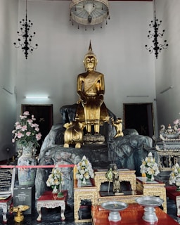 A large golden Buddha statue is the central focus, placed on a rocky pedestal. Surrounding the statue are various items, including ornate vases with pink and white flowers, smaller statues, incense holders, and decorative tables with intricate designs. The room features a high ceiling with hanging chandeliers and lamps, along with framed pictures or artworks on the walls.