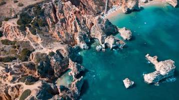An aerial view of a rugged coastline featuring craggy cliffs and turquoise waters. The cliffs are composed of reddish-brown rock formations, with green vegetation dotted across the landscape. Small beaches are nestled between the cliffs, with people visible on the sand. The clear blue-green water highlights several rock outcrops and small boats can be seen.