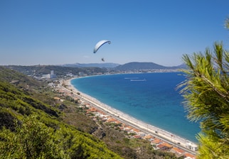 A paraglider soars above a beautiful coastal landscape with a sweeping view of the ocean. The coastline features a long, sandy beach bordered by green hills and dense vegetation. Houses are scattered along the shoreline, and a road runs parallel to the beach. Hills and distant mountains frame the background under a clear blue sky.