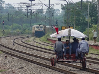 A manual rail trolley with four people seated on it is moving along the railway tracks. An umbrella is mounted on the trolley for shade, and a red flag is attached at the top. In the background, a train is approaching on a separate track. The scene is set in a rural or semi-urban environment with greenery surrounding the tracks. Electrical lines and railway infrastructure are visible overhead.