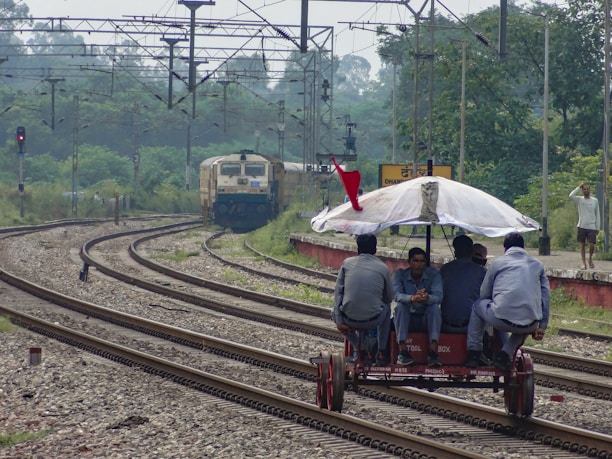 A manual rail trolley with four people seated on it is moving along the railway tracks. An umbrella is mounted on the trolley for shade, and a red flag is attached at the top. In the background, a train is approaching on a separate track. The scene is set in a rural or semi-urban environment with greenery surrounding the tracks. Electrical lines and railway infrastructure are visible overhead.