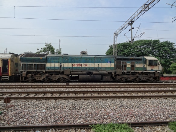 A diesel locomotive of the Indian Railways is stationary on a set of train tracks. The locomotive is bluish-green and beige in color, with visible text in a specific script on its side. Overhead power lines and an electric pole are present in the background. Trees and additional hardware infrastructure can also be seen, indicating a railway setting.