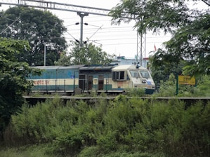 A train is moving along tracks surrounded by greenery and trees. Overhead power lines are visible, and there is a signboard with the name Dhandera on the platform.