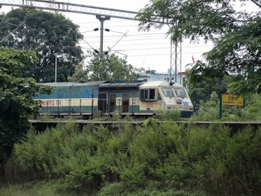 A train is moving along tracks surrounded by greenery and trees. Overhead power lines are visible, and there is a signboard with the name Dhandera on the platform.