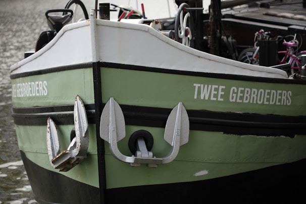 A green and white boat with the name 'TWEE GEBROEDERS' written on the side, featuring large metallic anchors attached to the bow. The background includes parts of the deck and some bicycles.