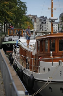 A man stands on the deck of a moored wooden boat, handling a hose. The boat is docked along a canal, lined with buildings and other boats. The background features trees with some yellowing leaves, and the scene appears to be set in an urban environment.