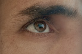 A close-up of a human eye displaying detailed textures of the skin, eyelashes, and the shape of the eyebrow. The eye is reflecting an image that seems to depict a person and some scenery, possibly a building or room.