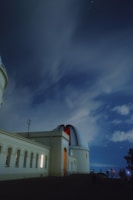 An observatory building with a large dome sits under a night sky filled with stars. The structure is illuminated by artificial lights, casting a warm glow on its pale walls. Silhouettes of several people are visible near the base, likely setting up telescopes or cameras. Wispy clouds drift across the sky, partially obscured by the long-exposure effect.