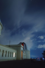 An observatory building with a large dome sits under a night sky filled with stars. The structure is illuminated by artificial lights, casting a warm glow on its pale walls. Silhouettes of several people are visible near the base, likely setting up telescopes or cameras. Wispy clouds drift across the sky, partially obscured by the long-exposure effect.