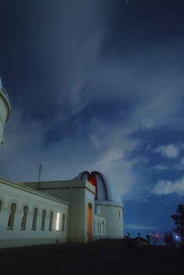 An observatory building with a large dome sits under a night sky filled with stars. The structure is illuminated by artificial lights, casting a warm glow on its pale walls. Silhouettes of several people are visible near the base, likely setting up telescopes or cameras. Wispy clouds drift across the sky, partially obscured by the long-exposure effect.