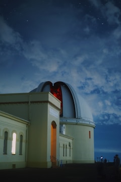 An observatory with a large dome partially open reveals the telescope inside. The building has an illuminated doorway and windows, set against a cloudy night sky with visible stars. A person stands outside looking at the structure.