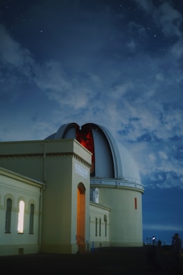 An observatory with a large dome partially open reveals the telescope inside. The building has an illuminated doorway and windows, set against a cloudy night sky with visible stars. A person stands outside looking at the structure.