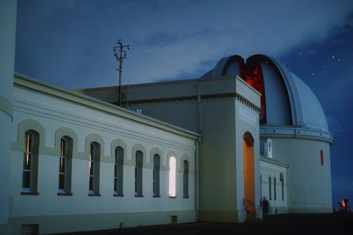 An observatory building with an open dome is captured under a night sky filled with stars. The structure features numerous windows along its side and is illuminated by a warm interior light. A person is visible near the entrance, which is slightly ajar, allowing a glimpse of the red-lit interior. The architecture is complemented by antennas and other equipment mounted on the roof.
