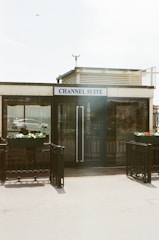 A small building with a sign reading 'Channel Suite' is prominently placed. The facade features large glass doors flanked by planters with colorful flowers. There is a metallic railing surrounding the entrance, and a bird is visible in the sky overhead.