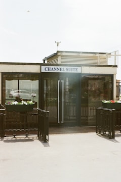 A small building with a sign reading 'Channel Suite' is prominently placed. The facade features large glass doors flanked by planters with colorful flowers. There is a metallic railing surrounding the entrance, and a bird is visible in the sky overhead.