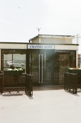 A small building with a sign reading 'Channel Suite' is prominently placed. The facade features large glass doors flanked by planters with colorful flowers. There is a metallic railing surrounding the entrance, and a bird is visible in the sky overhead.
