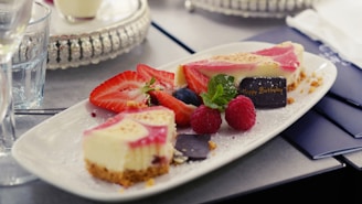 A rectangular white plate holds a selection of desserts, including a slice of cheesecake with a pink topping, fresh strawberries, raspberries, a blueberry, and a sprig of mint. A small chocolate piece with 'Happy Birthday' written on it in gold is also visible. The desserts are garnished with a dusting of powdered sugar. The background features a reflective surface and a glass nearby.