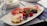 A rectangular white plate holds a selection of desserts, including a slice of cheesecake with a pink topping, fresh strawberries, raspberries, a blueberry, and a sprig of mint. A small chocolate piece with 'Happy Birthday' written on it in gold is also visible. The desserts are garnished with a dusting of powdered sugar. The background features a reflective surface and a glass nearby.