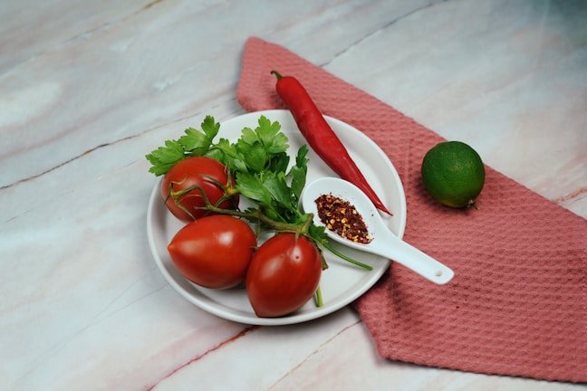 A white plate holds three ripe tomatoes with stems, accompanied by fresh green parsley. A bright red chili pepper is placed nearby, along with a white spoon filled with red pepper flakes. A whole lime rests on a textured pink cloth beside the plate, highlighting the fresh ingredients.