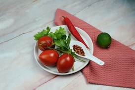 A white plate holds three ripe tomatoes with stems, accompanied by fresh green parsley. A bright red chili pepper is placed nearby, along with a white spoon filled with red pepper flakes. A whole lime rests on a textured pink cloth beside the plate, highlighting the fresh ingredients.