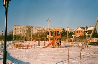 An outdoor playground in a residential area during winter. The playground equipment is brightly colored with red, yellow, and orange structures, including slides and climbing frames. The ground is covered in a light layer of snow. In the background, there is a large apartment building and a couple of houses. A lamppost and utility poles are also visible.