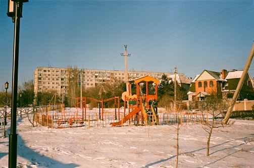 An outdoor playground in a residential area during winter. The playground equipment is brightly colored with red, yellow, and orange structures, including slides and climbing frames. The ground is covered in a light layer of snow. In the background, there is a large apartment building and a couple of houses. A lamppost and utility poles are also visible.