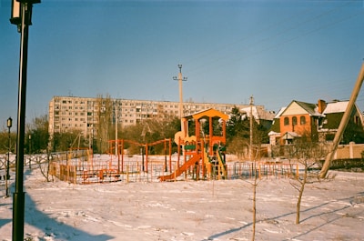 An outdoor playground in a residential area during winter. The playground equipment is brightly colored with red, yellow, and orange structures, including slides and climbing frames. The ground is covered in a light layer of snow. In the background, there is a large apartment building and a couple of houses. A lamppost and utility poles are also visible.