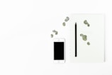 A minimalist arrangement featuring a white smartphone with a blank screen, a black pencil, and a light-colored notebook with scattered green leaves. The objects are placed on a clean, white background, emphasizing simplicity and elegance.