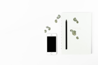 A minimalist arrangement featuring a white smartphone with a blank screen, a black pencil, and a light-colored notebook with scattered green leaves. The objects are placed on a clean, white background, emphasizing simplicity and elegance.
