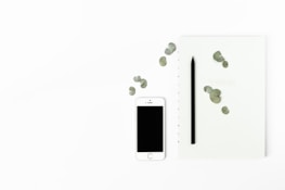A minimalist arrangement featuring a white smartphone with a blank screen, a black pencil, and a light-colored notebook with scattered green leaves. The objects are placed on a clean, white background, emphasizing simplicity and elegance.