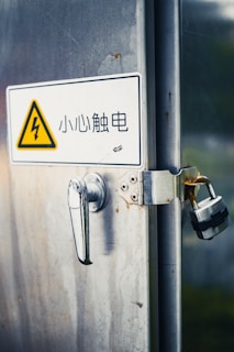 A metal door with rust spots features a handle and a padlock securing it. A caution sign with an electrical warning symbol is displayed, accompanied by Chinese text.