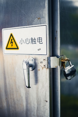 A metal door with rust spots features a handle and a padlock securing it. A caution sign with an electrical warning symbol is displayed, accompanied by Chinese text.