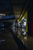 A shopping mall corridor is shown with dim lighting, reflective polished floors, and modern architectural elements. On the right side, a brightly lit storefront labeled 'POP MART' stands out, attracting some people inside. The ceiling features recessed lighting and architectural curves, while reflections of lights on the floor add a visual depth.