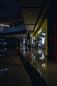 A shopping mall corridor is shown with dim lighting, reflective polished floors, and modern architectural elements. On the right side, a brightly lit storefront labeled 'POP MART' stands out, attracting some people inside. The ceiling features recessed lighting and architectural curves, while reflections of lights on the floor add a visual depth.