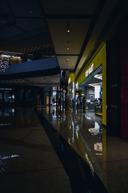 A shopping mall corridor is shown with dim lighting, reflective polished floors, and modern architectural elements. On the right side, a brightly lit storefront labeled 'POP MART' stands out, attracting some people inside. The ceiling features recessed lighting and architectural curves, while reflections of lights on the floor add a visual depth.