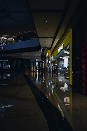 A shopping mall corridor is shown with dim lighting, reflective polished floors, and modern architectural elements. On the right side, a brightly lit storefront labeled 'POP MART' stands out, attracting some people inside. The ceiling features recessed lighting and architectural curves, while reflections of lights on the floor add a visual depth.