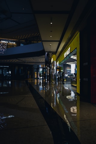 A shopping mall corridor is shown with dim lighting, reflective polished floors, and modern architectural elements. On the right side, a brightly lit storefront labeled 'POP MART' stands out, attracting some people inside. The ceiling features recessed lighting and architectural curves, while reflections of lights on the floor add a visual depth.