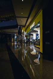 A modern shopping mall corridor with a highly polished floor that reflects the overhead lights and store displays. The POP MART store is prominently visible with a bright yellow facade and glowing signage. People are seen walking near the store, adding to the lively atmosphere.