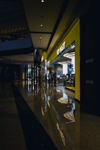 A modern shopping mall corridor with a highly polished floor that reflects the overhead lights and store displays. The POP MART store is prominently visible with a bright yellow facade and glowing signage. People are seen walking near the store, adding to the lively atmosphere.