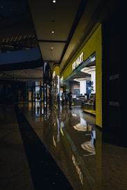 A modern shopping mall corridor with a highly polished floor that reflects the overhead lights and store displays. The POP MART store is prominently visible with a bright yellow facade and glowing signage. People are seen walking near the store, adding to the lively atmosphere.