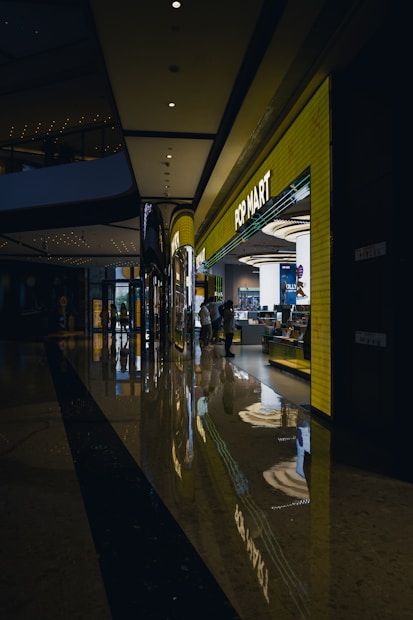A modern shopping mall corridor with a highly polished floor that reflects the overhead lights and store displays. The POP MART store is prominently visible with a bright yellow facade and glowing signage. People are seen walking near the store, adding to the lively atmosphere.