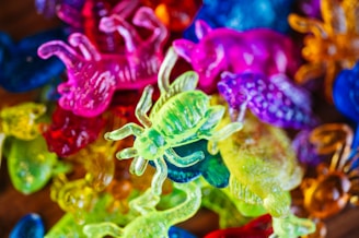 A collection of colorful, translucent plastic toy bugs and animals. The toys are piled together in various vibrant hues, including green, pink, yellow, blue, orange, and purple. The details of the textures and shapes of the toys are clearly visible.