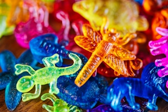 A vibrant assortment of colorful, translucent plastic animal toys including scorpions, lizards, and insects. The toys overlap and are lit with bright lighting, enhancing the vivid colors and glossy texture.