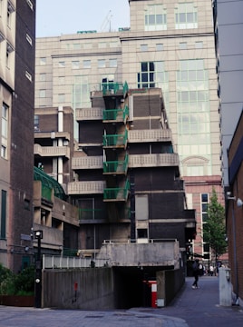 A modern urban building complex featuring a combination of brick and glass. The structure exhibits an intricate design with a series of protruding balconies accented by green railings. The surrounding area includes adjacent buildings and a paved walkway, with scattered greenery and a few people walking.