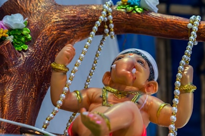 A detailed statue of a young deity with an elephant head, adorned with gold jewelry and sitting on a swing. The swing is decorated with pearl-like beads and is attached to a branch with vibrant flowers. The deity is depicted as playful and innocent.