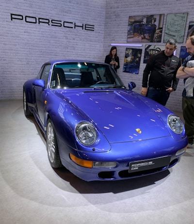 A blue Porsche sports car is on display in what appears to be a showroom or exhibit space. The car is parked on a light-colored floor against a backdrop of white brick walls, adorned with the Porsche logo and several framed pictures. There are three people nearby, with two of them looking at the car and one using a mobile device.