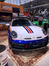 A sleek, white and blue car with red and yellow stripes is displayed indoors. It features a robust design and is parked on an ornate carpet. The environment suggests an exhibition or auto show, with people gathered around and informational displays visible in the background.