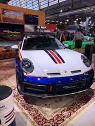 A sleek, white and blue car with red and yellow stripes is displayed indoors. It features a robust design and is parked on an ornate carpet. The environment suggests an exhibition or auto show, with people gathered around and informational displays visible in the background.