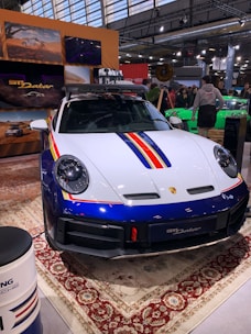 A sleek, white and blue car with red and yellow stripes is displayed indoors. It features a robust design and is parked on an ornate carpet. The environment suggests an exhibition or auto show, with people gathered around and informational displays visible in the background.