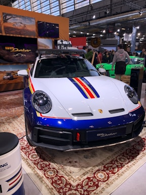 A sleek, white and blue car with red and yellow stripes is displayed indoors. It features a robust design and is parked on an ornate carpet. The environment suggests an exhibition or auto show, with people gathered around and informational displays visible in the background.